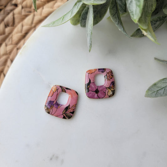 Two floral-patterned square earrings on a white surface with green leaves in the background.
