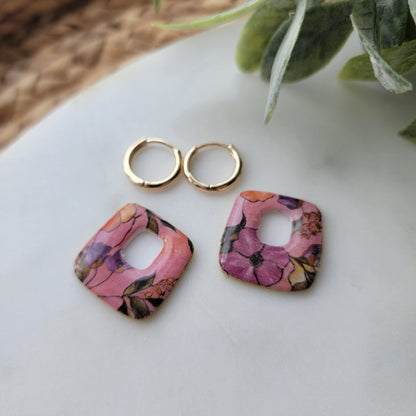 Two floral-patterned earrings and two gold rings on a white surface with a plant in the corner.
