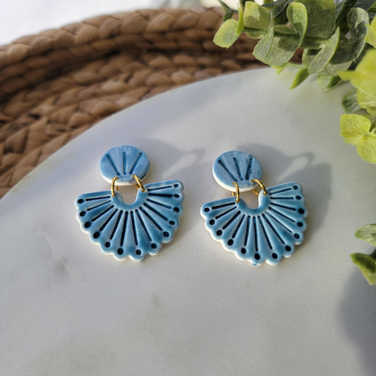 Blue earrings with gold hooks on a white surface with green leaves in the background