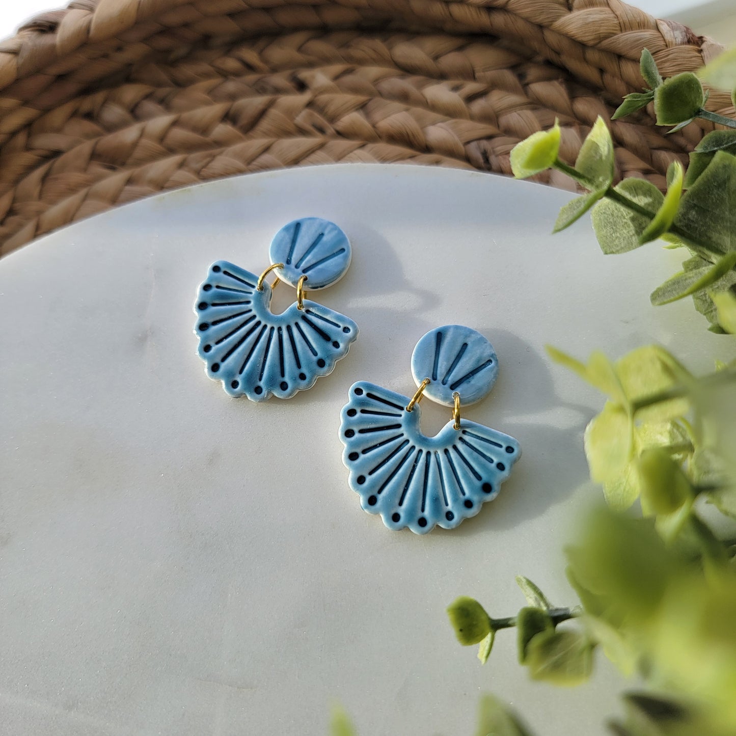 Blue ceramic earrings on a white surface with green leaves in the background