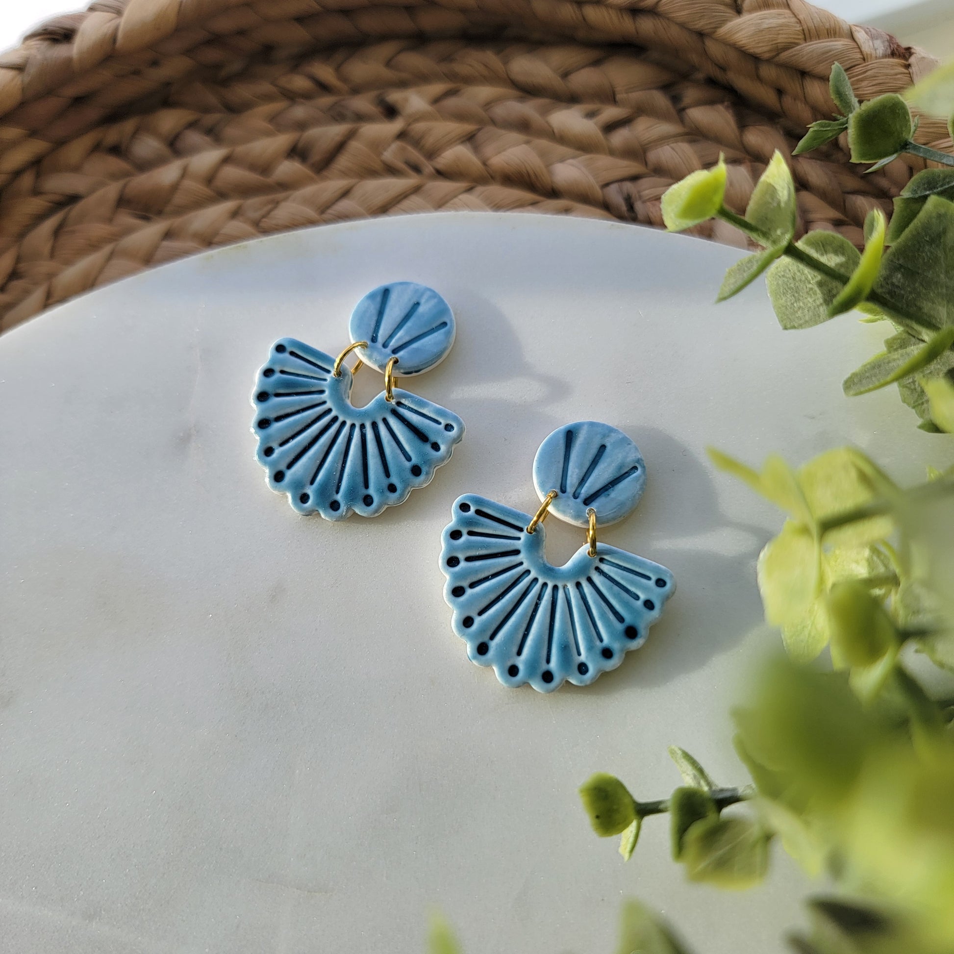 Blue ceramic earrings on a white surface with green leaves in the background