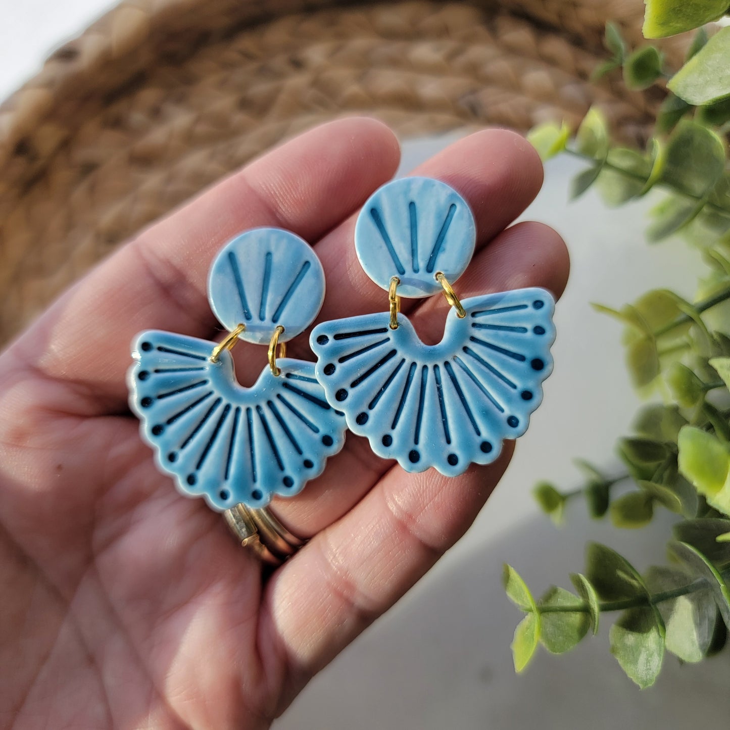 Hand holding blue fan-shaped earrings with a blurred green plant in the background