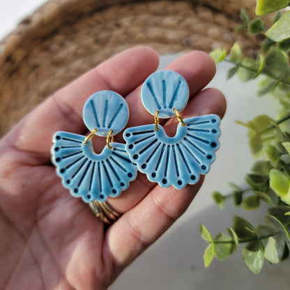 Hand holding blue fan-shaped earrings with a blurred green plant in the background