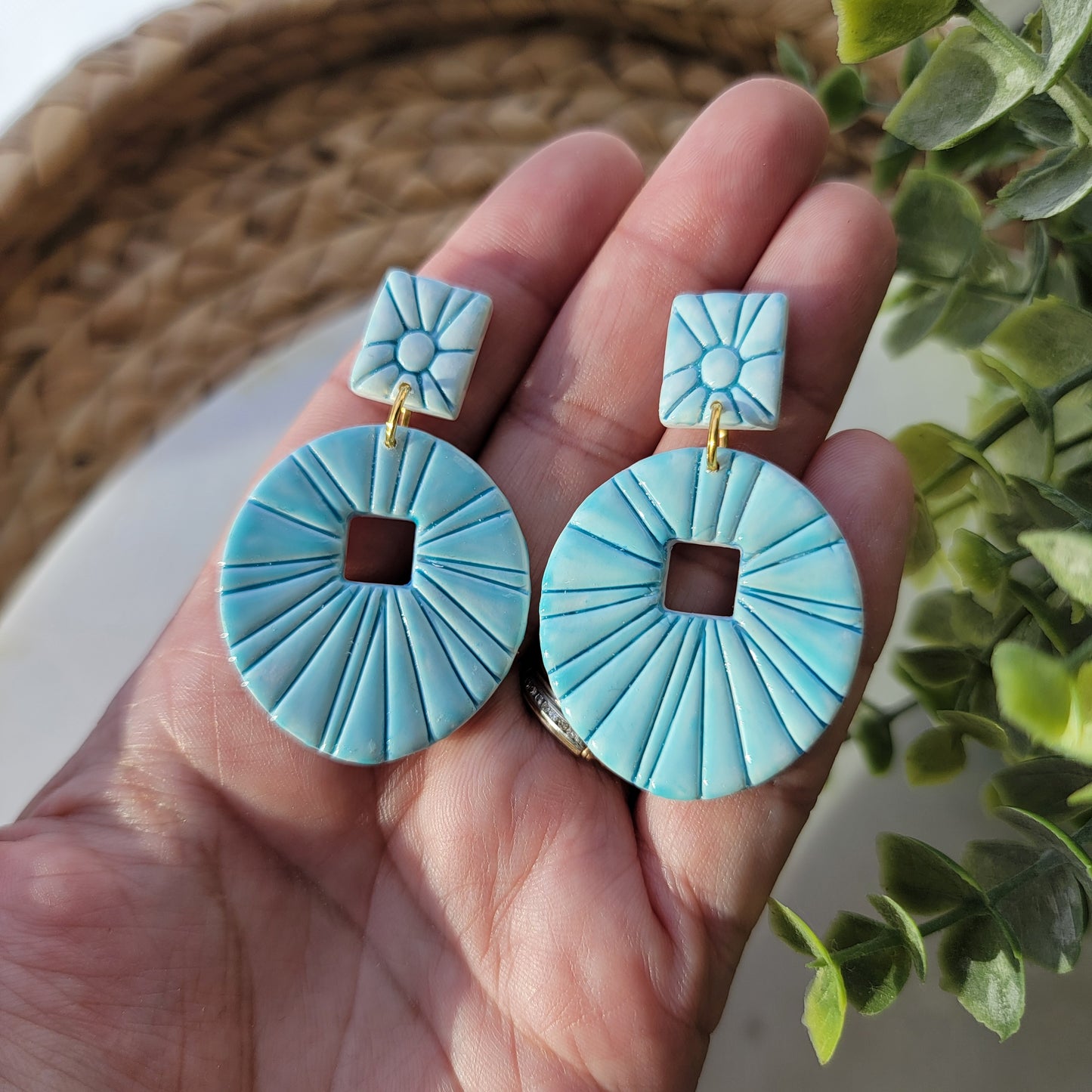 Pair of teal earrings held in a hand on a marble surface with green leaves in the background