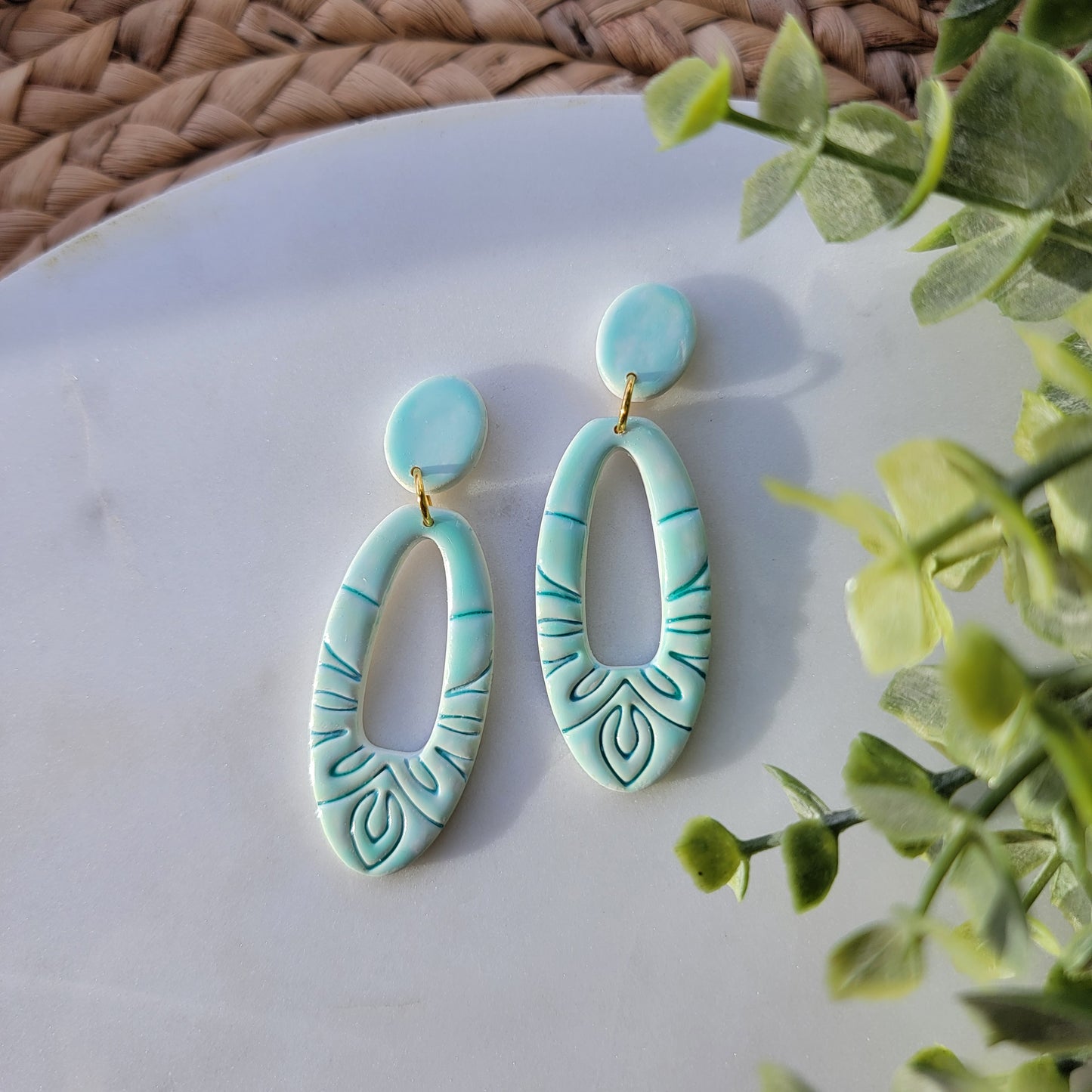 Turquoise earrings with leaf patterns on a white surface with green leaves in the background