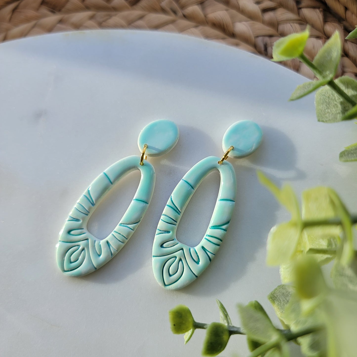 Turquoise earrings with leaf patterns on a white surface with green leaves in the background