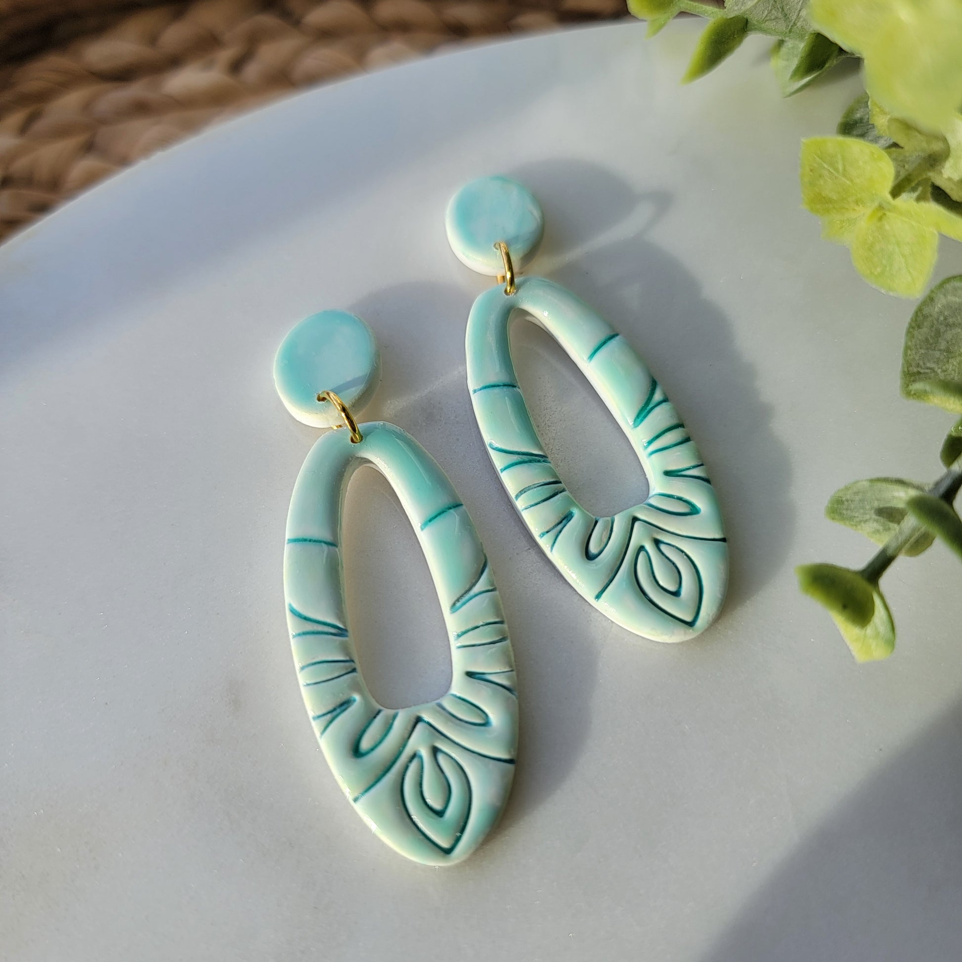 Turquoise earrings with leaf patterns on a white surface with green leaves in the background