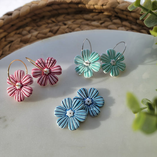 Flower-shaped earrings in green, blue, and pink on a white surface with a woven mat.