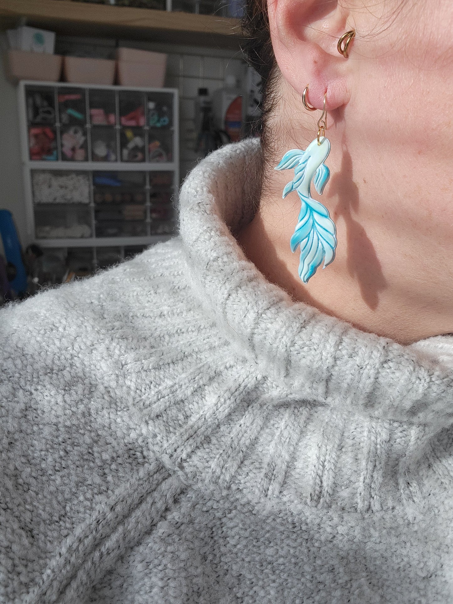 Person wearing a gray sweater with a koi fish shaped earring in the colors of blue and green, showing sizing of earrings.