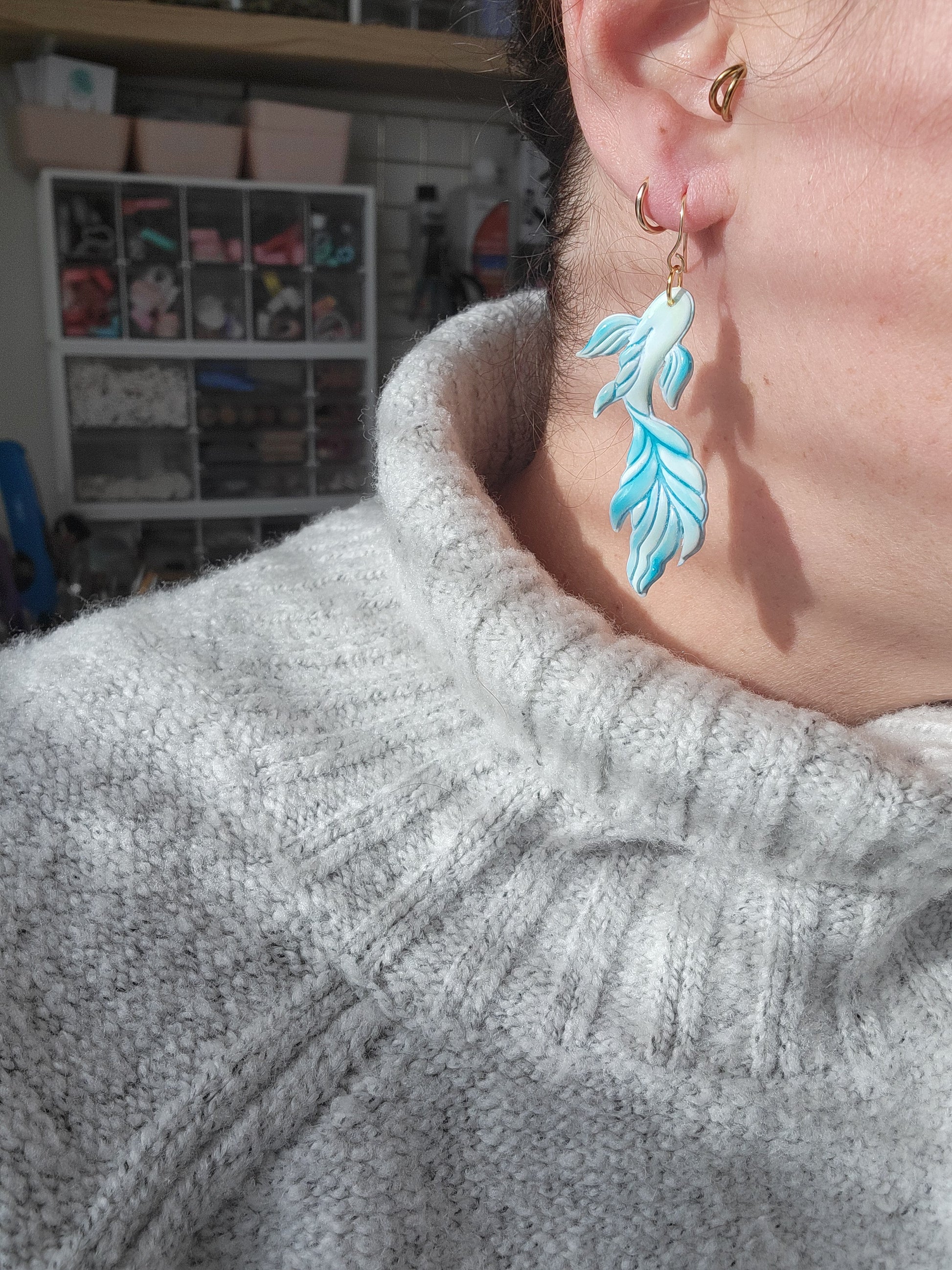 Person wearing a gray sweater with a koi fish shaped earring in the colors of blue and green, showing sizing of earrings.