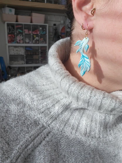 Person wearing a gray sweater with a koi fish shaped earring in the colors of blue and green, showing sizing of earrings.