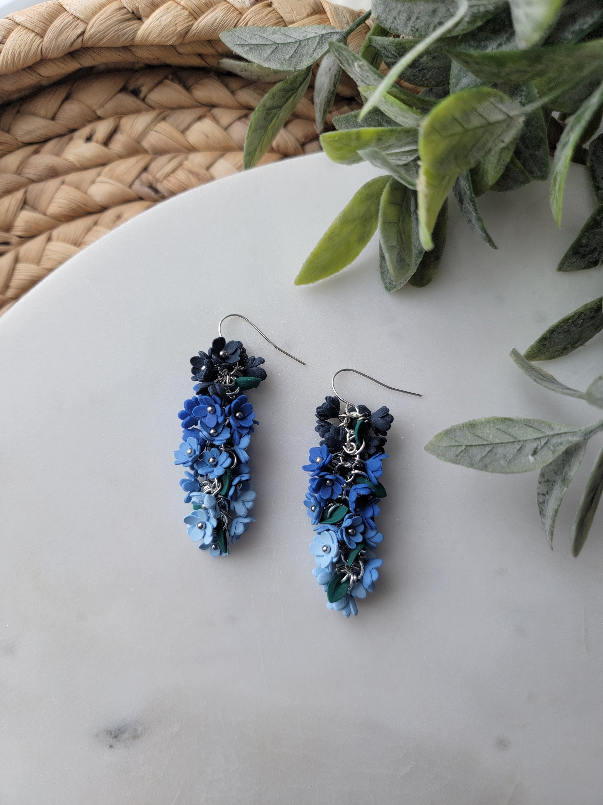 Pair of blue floral earrings on a white surface with green leaves in the background