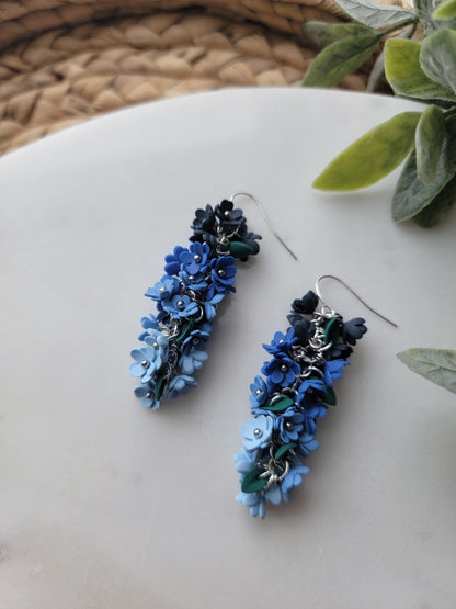 Blue floral earrings on a light surface with green leaves in the corner.