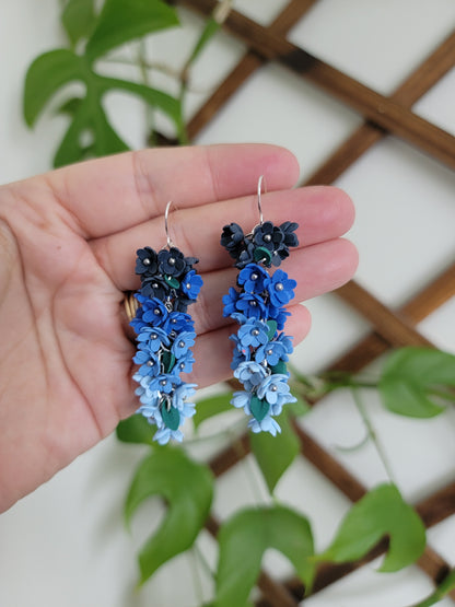 Blue floral earrings held in a hand with a green leafy background