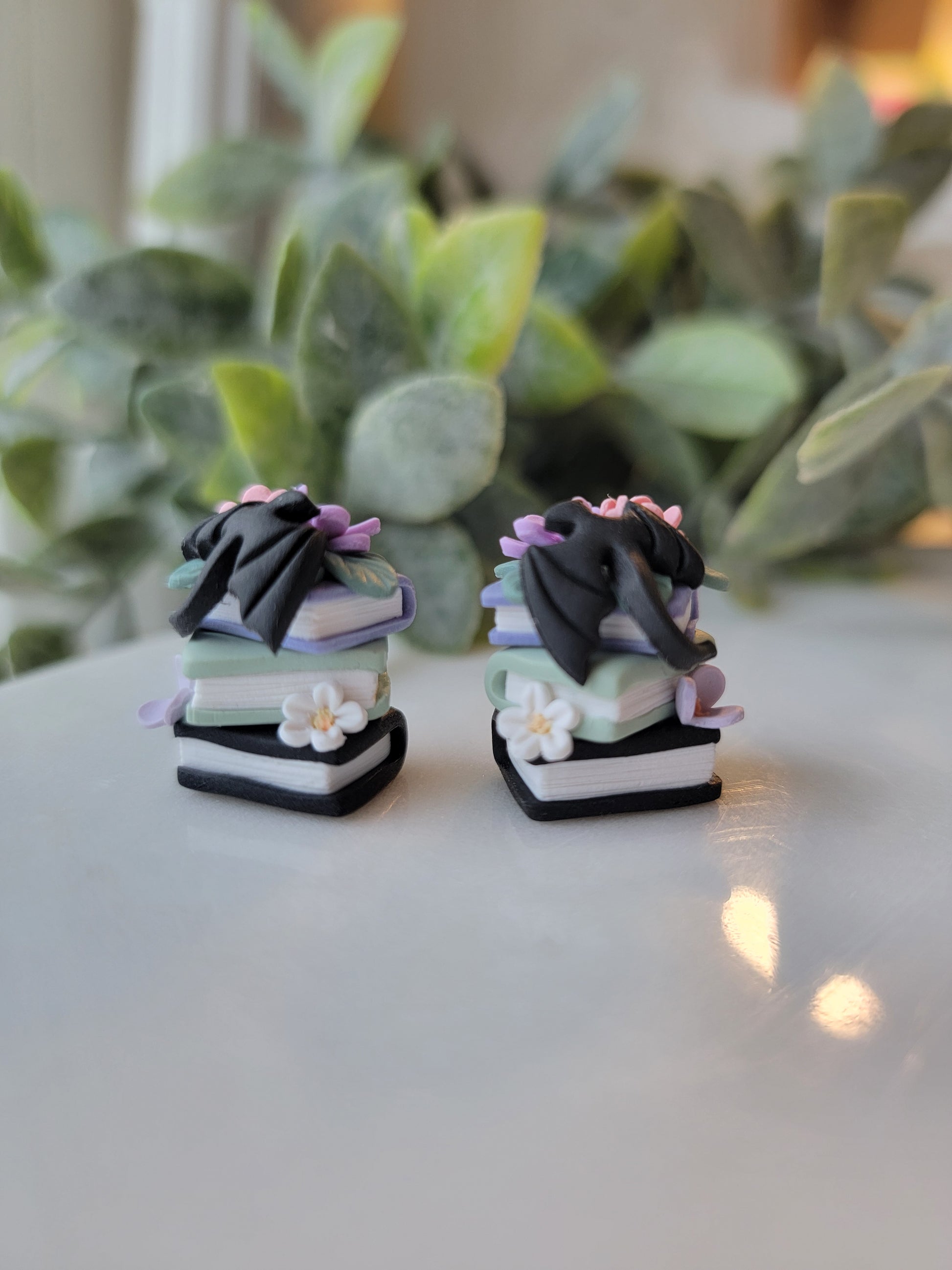 A set of earrings featuring colorful book stacks with dainty flowers and a black dragon on them.