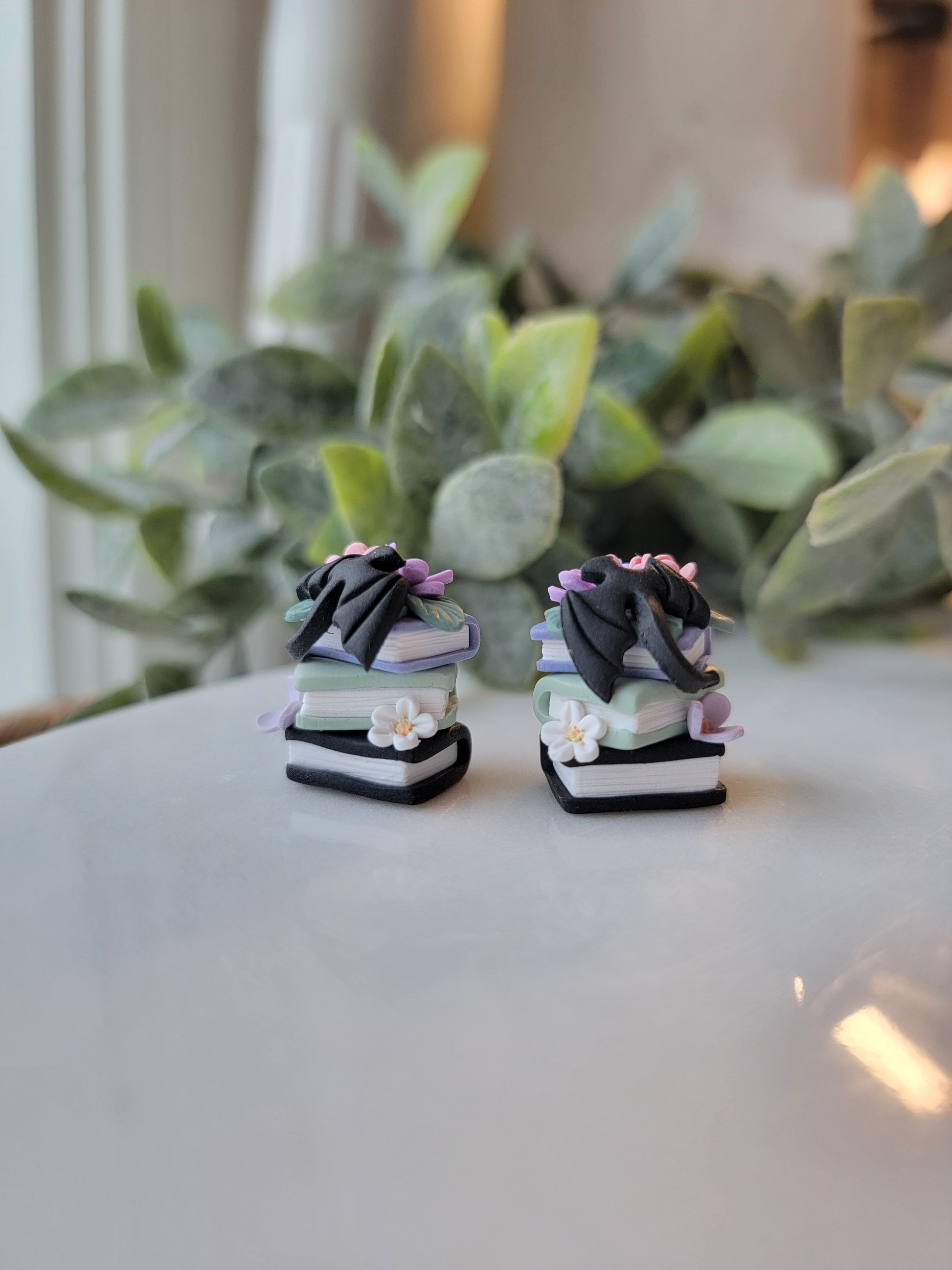 A set of earrings featuring colorful book stacks with dainty flowers and a black dragon on them.