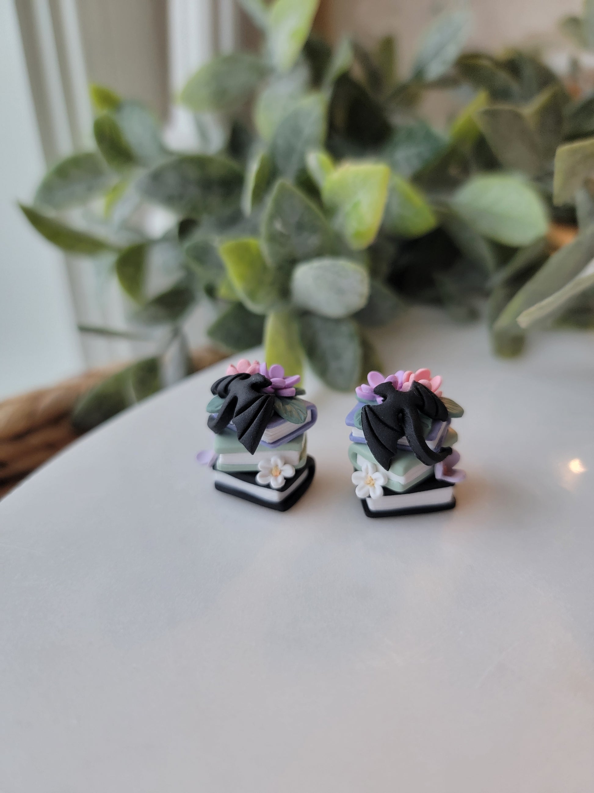 A set of earrings featuring colorful book stacks with dainty flowers and a black dragon on them.